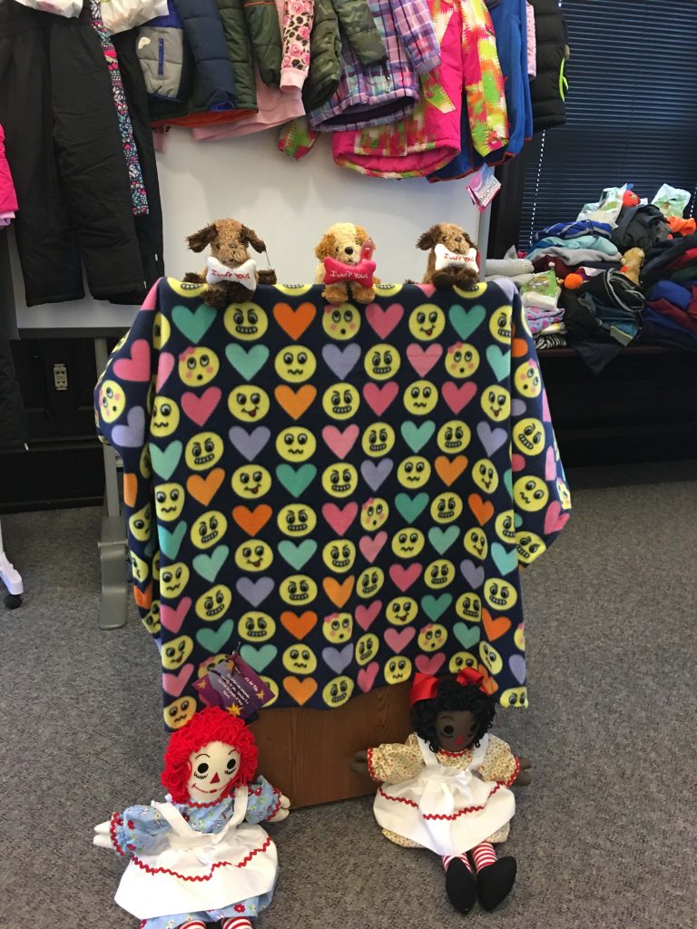 Winter coats, stuffed toy dogs and rag dolls placed near a black cloth with pastel hearts, various smiley face emojis prints draped over a wooden podium.