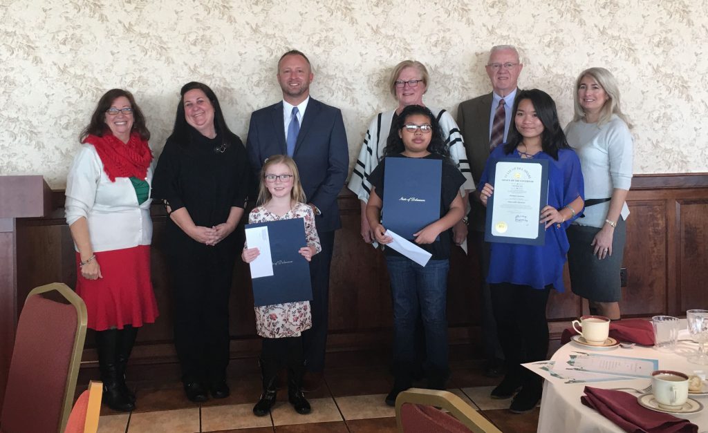 Young 2017 Poster Contest Award recipients standing with certificates standing in front of several adults and GACEC staff in front of a light colored wall with dark wood trim on the bottom of the wall.