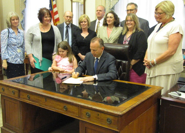 Seated at a clear reflective desk with a child next to Governor Jack Markel signing the Restraints and Seclusion Bill with GACE staff, elected officials and others standing behind and on the side of him.